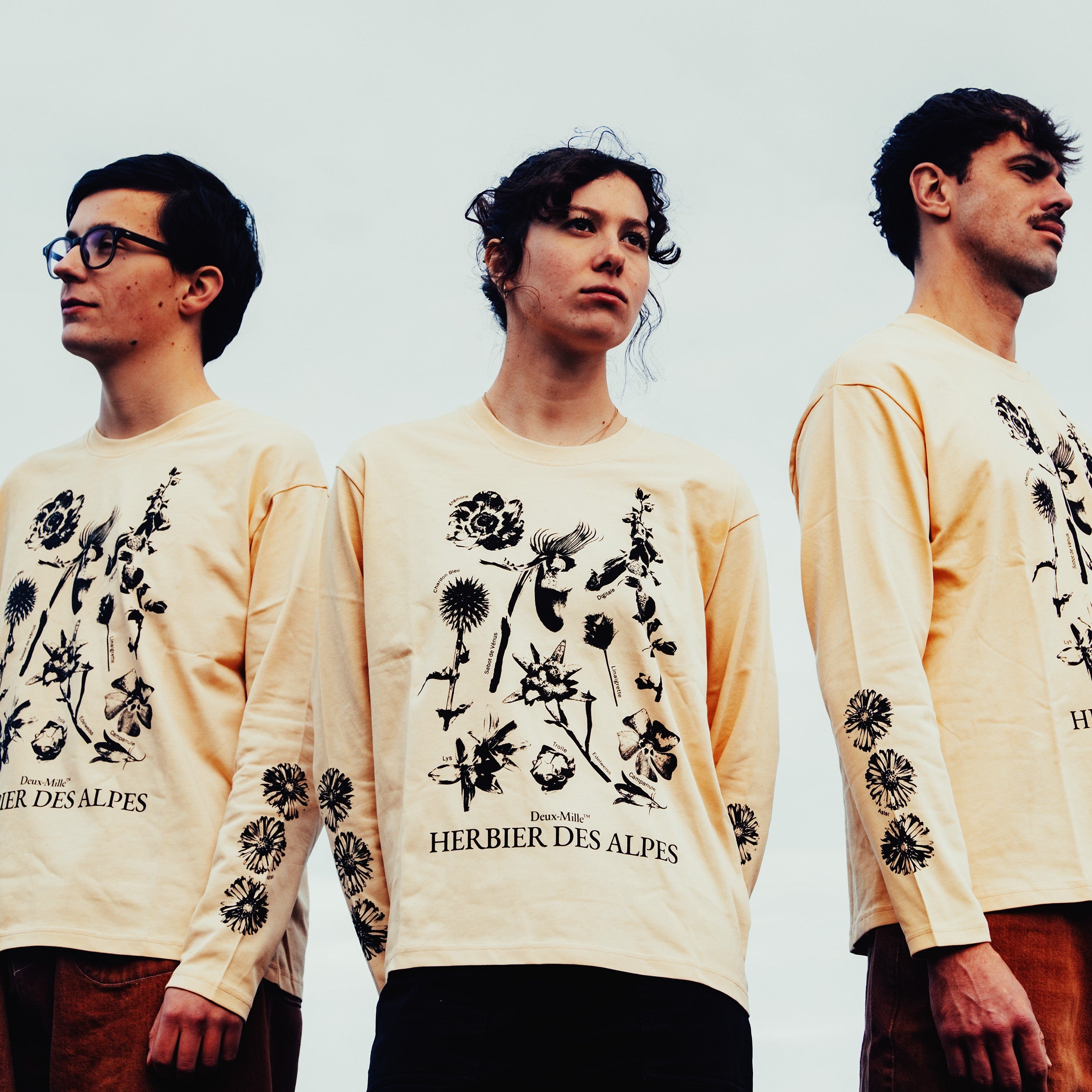 Three people wearing matching beige "Herbier des Alpes" long-sleeve shirts with black floral patterns on a light gray background